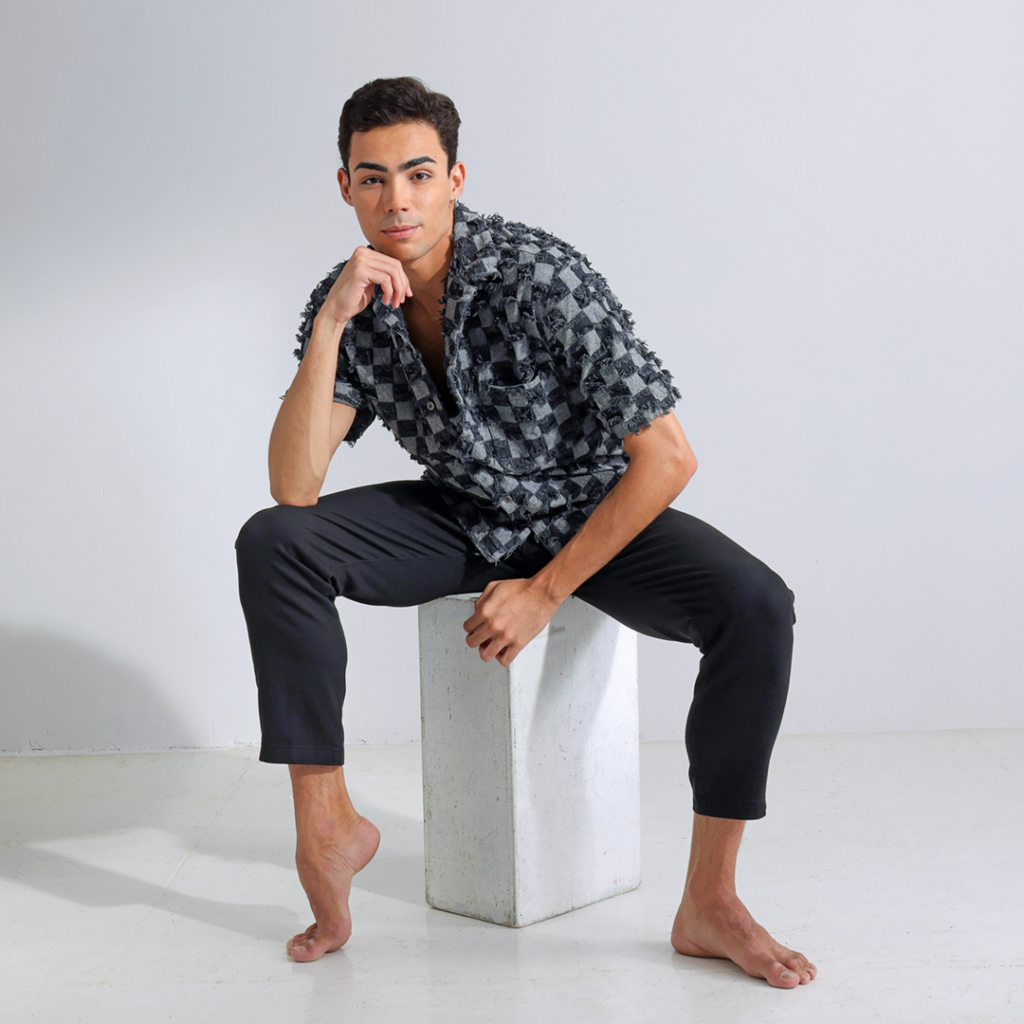 Victor Caixeta sits on a white rectangular box in front of a white backdrop. He rests his right elbow onto his right knee and leans his chin onto his right hand. His right foot is popped, showing offf his high arches. He wears a gray and black checkered shirt and black pants, and looks directly into the camera with a calm smile.