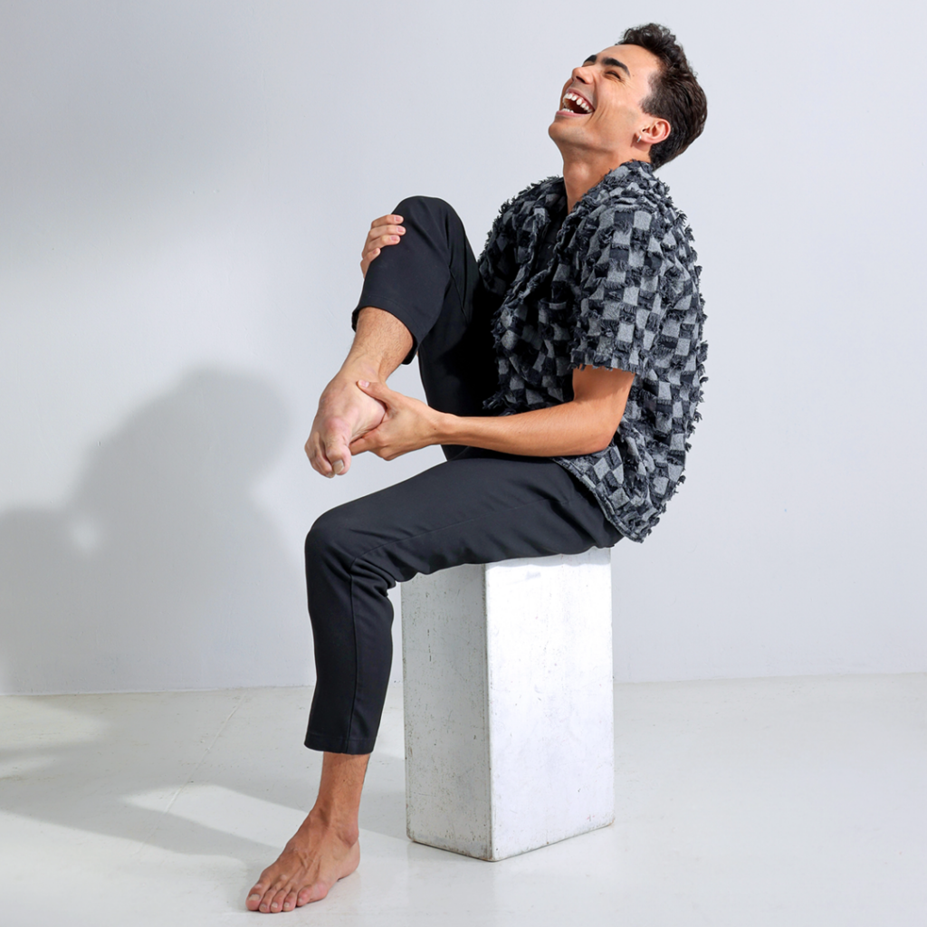 Victor Caixeta sits on a white rectangular box in front of a white backdrop. His body is angled to his right so that he is slightly in profile. He throws his head back and laughs and he lifts his right knee up, holding onto his pointed right foot. He wears a gray and black checkered shirt and black pants.