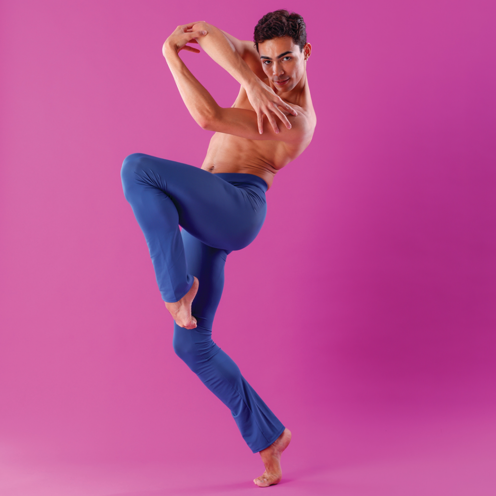 Victor Caixeta, wearing blue yoga pants, is shown slightly in profile facing his right. He lifts his left leg into a parallel retiré and relevés onto a bent right leg. He crosses his arms, leaving space between them and his body, and looks intensely into the camera.