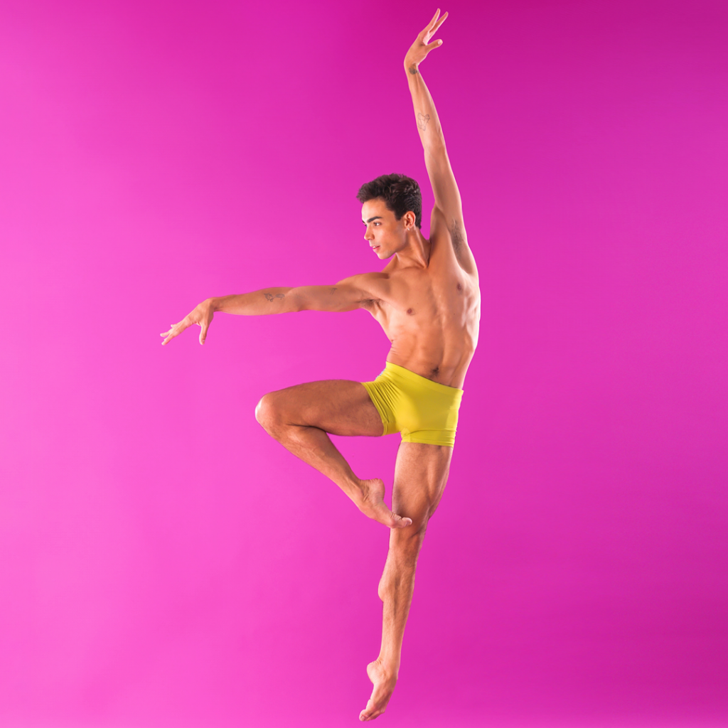 Victor Caixeta is shown jumping in front of a hot pink backdrop. He is shirtless and barefoot and wears yellow shorts. He jumps off of his left leg with his right leg in a retiree position, and reaches his right arm out to the side and his left arm straight up alongside his head. He looks intensely out over his right hand.