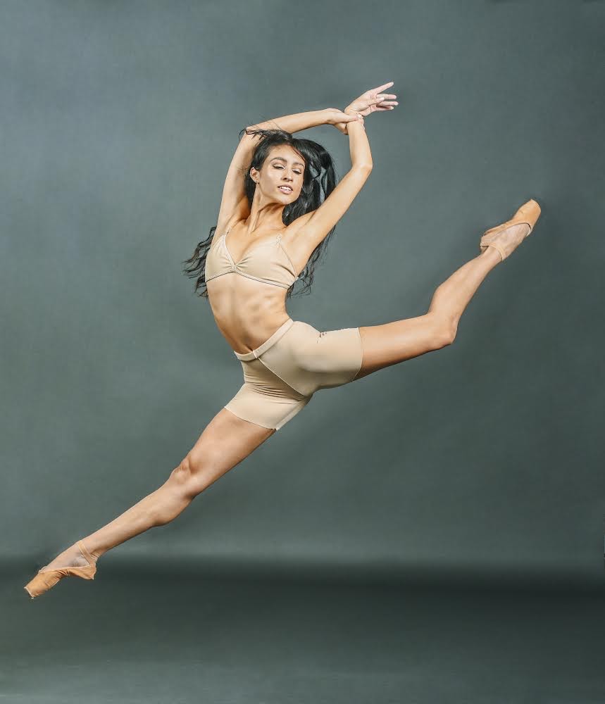 Paola McCormick Quintero jumps up nto a sissone with her left leg in attitude and her arms above her head. She grabs her left wrist with her right hand and looks out and down to the left. She wears a tan bra and bike shorts and tan pointe shoes, and jumps in front of a gray backdrop.