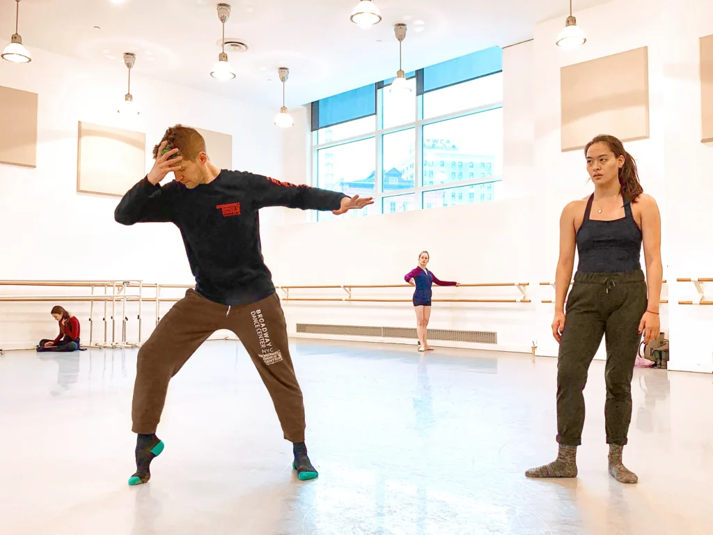 Barry Kerollis shows dancer Serena Lu a dance move during a rehearsal in a large, airy dance studio space. Wearing a dark long-sleeved t-shirt, olive-colored sweatpants and gray socks, he props his right foot onto demi-pointe and leans forward slightly, holding his head in his right hand as he stretches his left arm back. Lu, in a dark leotard, sweatpants, and socks, stands to his left and watches.