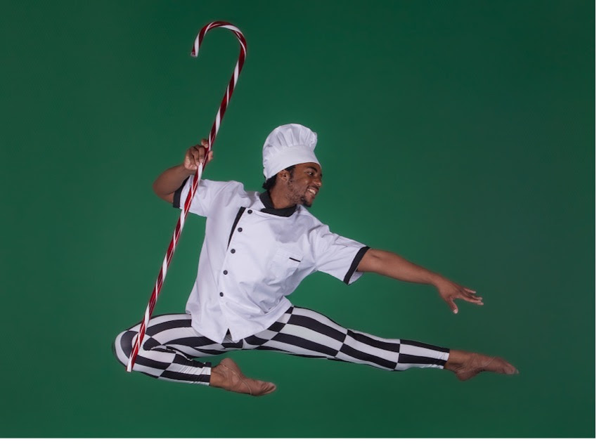 Robert Taylor Jr. does an Italian pas de chat, dressed as a Polar Express chef. He wears striped black and white pants, a white frock, and a white chef's hat while holding a large candy cane. He poses in front of a dark green backdrop.