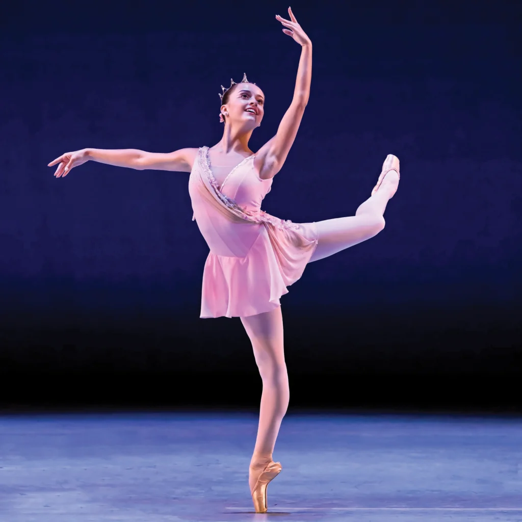 Sylvie Squires does a piqué attitude derriere on pointe during a performance at a ballet competition. She lifts her left leg in attitude effacé, with her left arm up and her right arm out to the side. She looks up toward her left hand and smiles brightly. Squires wear a short pink dance dress with a diagonal silver embellishment, a crown, pink tights and pink pointe shoes.