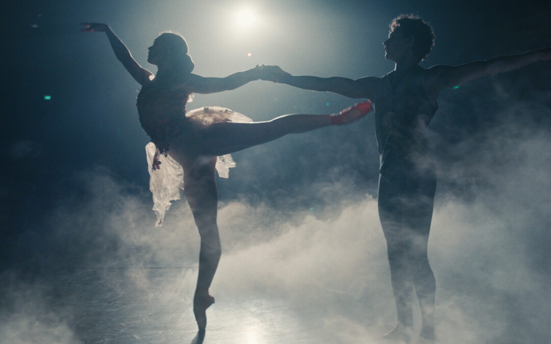 In a still from The Red Shoes: Next Step, the silhouettes of a female and male dancer are shown onstage, surrounded by dry ice. As she piques forward on pointe in arabesque, he reaches for her back arm.