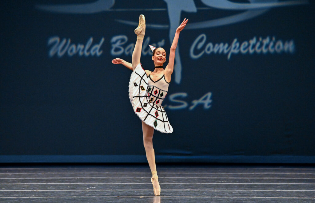 Ana Lorenzo performs a high développé relevé in effacé devant with her right leg during an onstage ballet competition. She holds her left arm high and her right amr out to the side and looks out to the audience with a large, confident smile. She waers a white tutu with multicolored diamond shapes on the skirt and bodice, a small party hat, a black choker neckalce, and pink tights and pointe shoes.