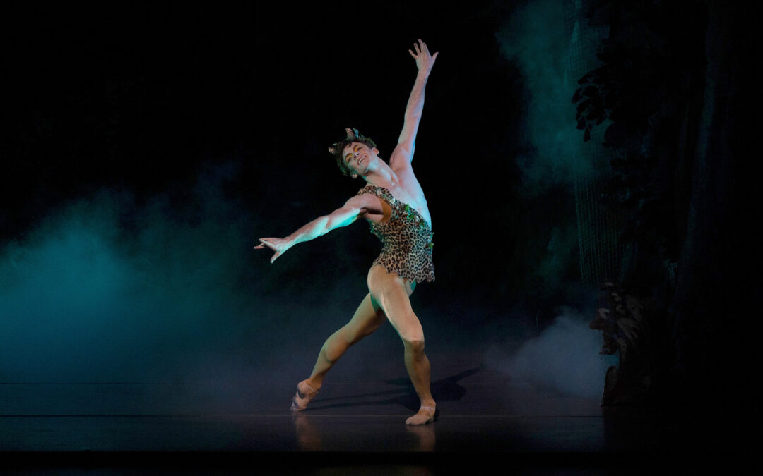 Jake Roxander dances onstage as Puck from Sir Frederick Ashton's The Dream. He poses in a croisé tendu derriere in plié, extending his arms wide. He wears a one-shouldered leopard print top with leaves trimming the edges, a leafy headpiece with two small horns, and mustard-colored tights and ballet slippers.