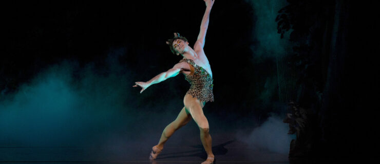 Jake Roxander dances onstage as Puck from Sir Frederick Ashton's The Dream. He poses in a croisé tendu derriere in plié, extending his arms wide. He wears a one-shouldered leopard print top with leaves trimming the edges, a leafy headpiece with two small horns, and mustard-colored tights and ballet slippers.