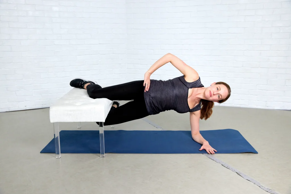 Eliza Tollett is demonstrating the third step of a Copenhagen plank. She is on a blue mat and wearing black pants, a gray top, black sneakers, and she is using a white bench.