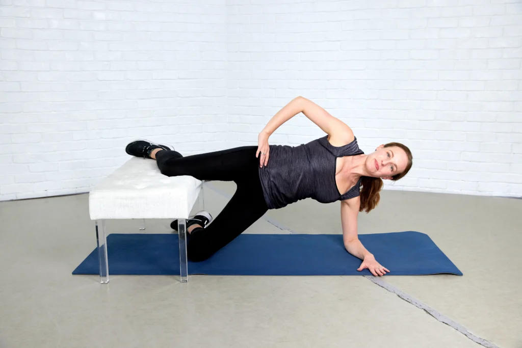 Eliza Tollett is demonstrating the second step of a Copenhagen plank. She is on a blue mat and wearing black pants, a gray top, black sneakers, and she is using a white bench.
