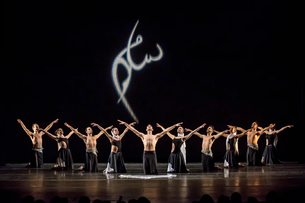 A group of 10 dancers kneel on the ground and create a V-shape formation, with a male dancers at the forefront center stage and two lines angling back on both side, alternating man-woman. They all extend their arms into a V-shape and lean slightly back on both knees. The men are shirtless and wear loose-fitting black pants, while the women wear black leotards with a cut-out on the midriff, long black skirts, and white scarves that wrap around their necks and attach at the back of their heads. They pose in front of a black backdrop with white Arabic lettering projected in the center.