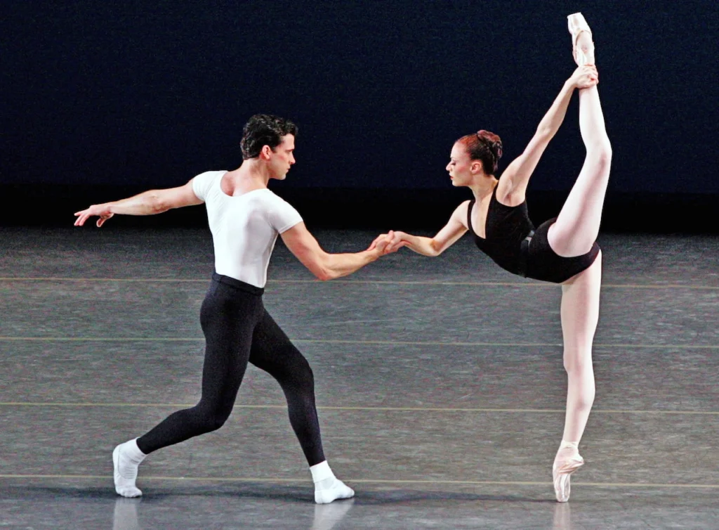 Carlos Guerra and Jennifer Kronenberg dance a pas de deux onstage. They face each other, and Kronenberg stands on pointe and pulls her left leg up into a high attitude over her head with her left hand. She holds Guerra's right hand with her own right hand as he pliés both legs and stretches his left arm out behind him. Kronenberg wears a blac leotard and pink tights, while Guerra wears a white t-shirt, black tights and white socks and ballet slippers.