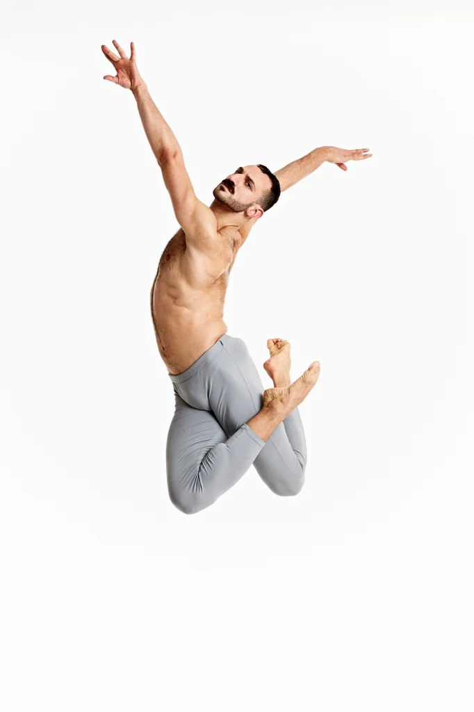 Fadi Khoury jumps up into the air and tucks his legs under him, crossing his right ankle over his left thigh. He reaches his arms up high, twists his torso so that his left arm is in front, and arches back, looking over his left shoulder. He wears gray tights and jumps in front of a white backdrop.