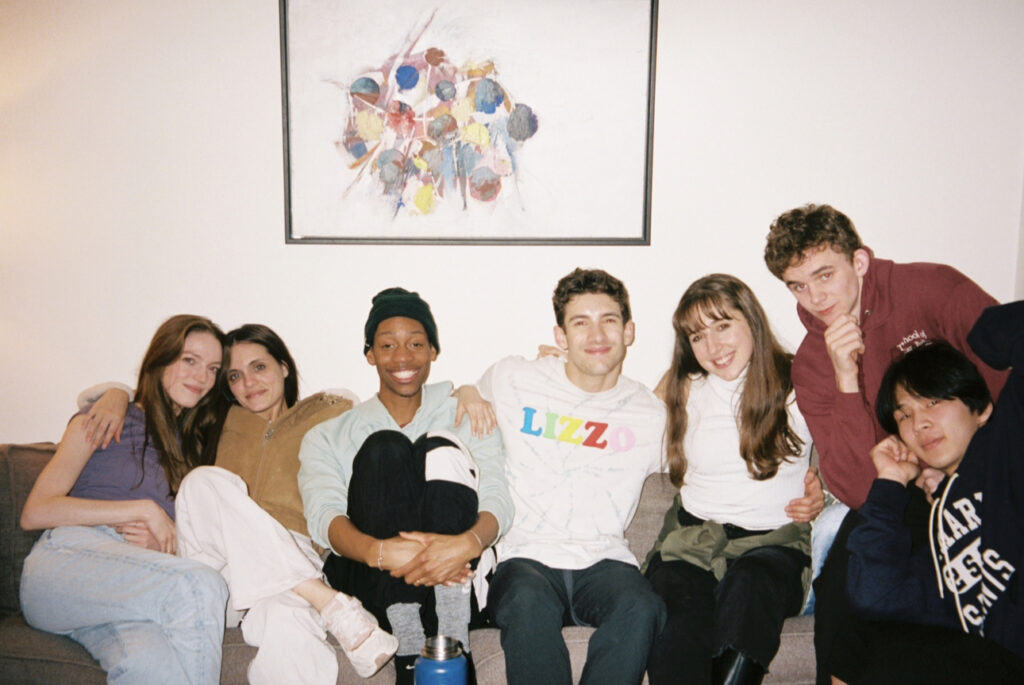 A group of seven people gather closely on a couch and smile for the camera. They are dressed casually and pose underneath a large print haning on the wall.