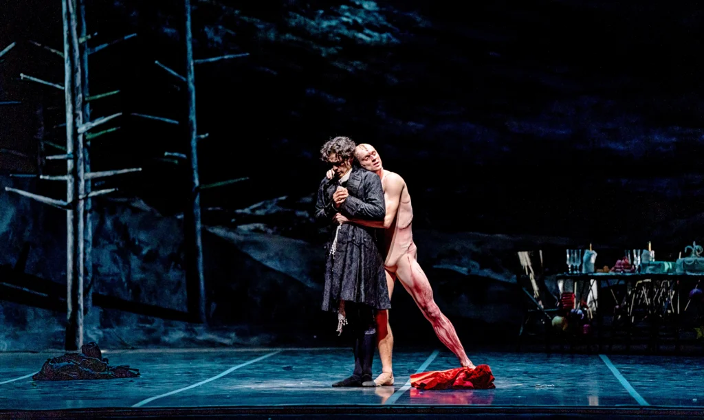 José Pablo Castro Cuevas, wearing a dark knee-length brocaded jacket, black tights and black ballet slippers, stands onstage facing the downstage right corner, clasping his hands and looking down pensively. Jonathan Dole, costumed in a nude unitard that appears bloodied and a nude skull-cap with a large scar going down the middle, hugs him from behind and closes his eyes. Behind them the set is dark with jagged, leafless trees and a table with various items on it.