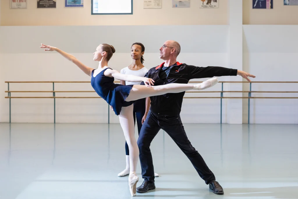 Stéphane Leonard, a ballet teacher, works with a male and female student in a large dance studio on some partnering moves. The girl, wearing a blue leotard and skirt and pink tights and shoes, does a first arabesque on pointe. Léonard—wearing a dark shirt, jeans and sneakers—stands behind her and lunges on his right leg, holding her hip with his right hand and reaching his left arm behind him. The boy, wearing a white t-shirt, black tights and white socks and ballet slippers, stands behind them and watches.