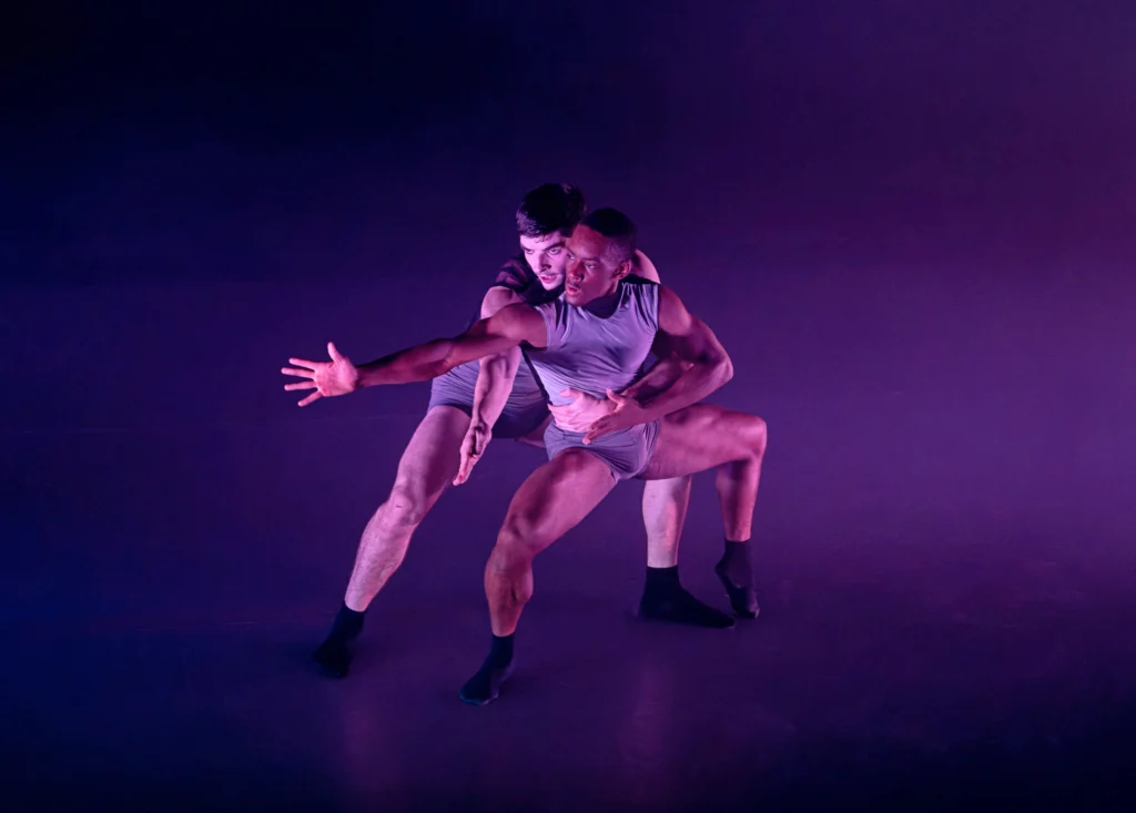 Two male dancers in dark gray biketards perform onstage, bathed in purple light. One dancer holds the waist of the other and pulls him close as they both plié in second position and stretch their right arm out to the side, their hands spread wide. The dancer in front opos his left foot up into demi-pointe, and they both look intently off towards stage right.
