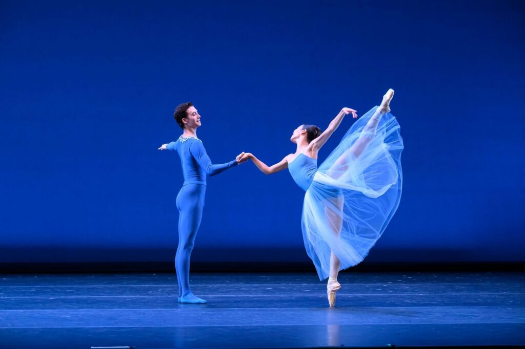 A male and female dancer perform "Serenade" onstage in deep blue costumes. He holds her hand as she does a huge arabesque facing him, her long gauzy skirt flying up behind her.