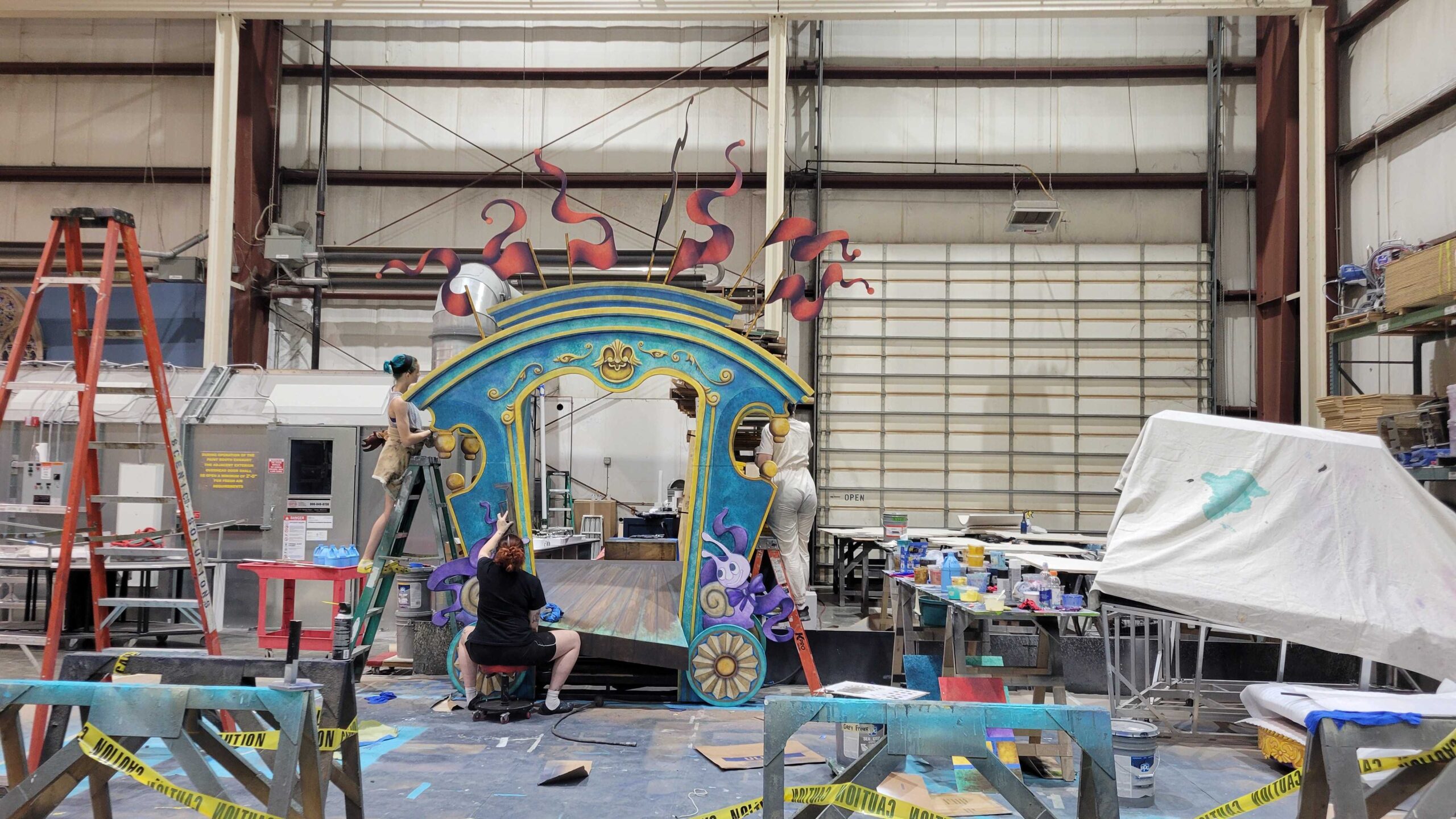 Backstage, stagehands and designers work on creating a large blue structure with pink decorative accents