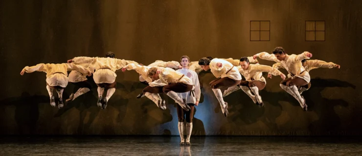 A male dancer stands center stage with his arms at his sides, looking directly out to the audience with a black expression. A corps of men create a circle around him and jump up into the air, legs tucked underneath them as they bend their upper bodies forward and lift their curved arms out to the side. All of the dancers wear white peasant blouses, bown knickers, white tights and brown ballet shoes. They dance in front of a brown backdrop with two windows sketched in black towards the stage left side.