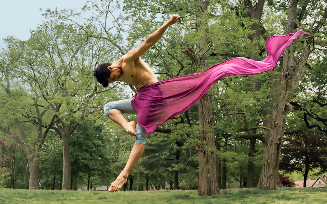 Manish Chauhan flies through the air outside in a stylized jump in retire devant, his body pitched forward and arms thrown back, a magenta scarf flowing behind him as it wraps around his front.