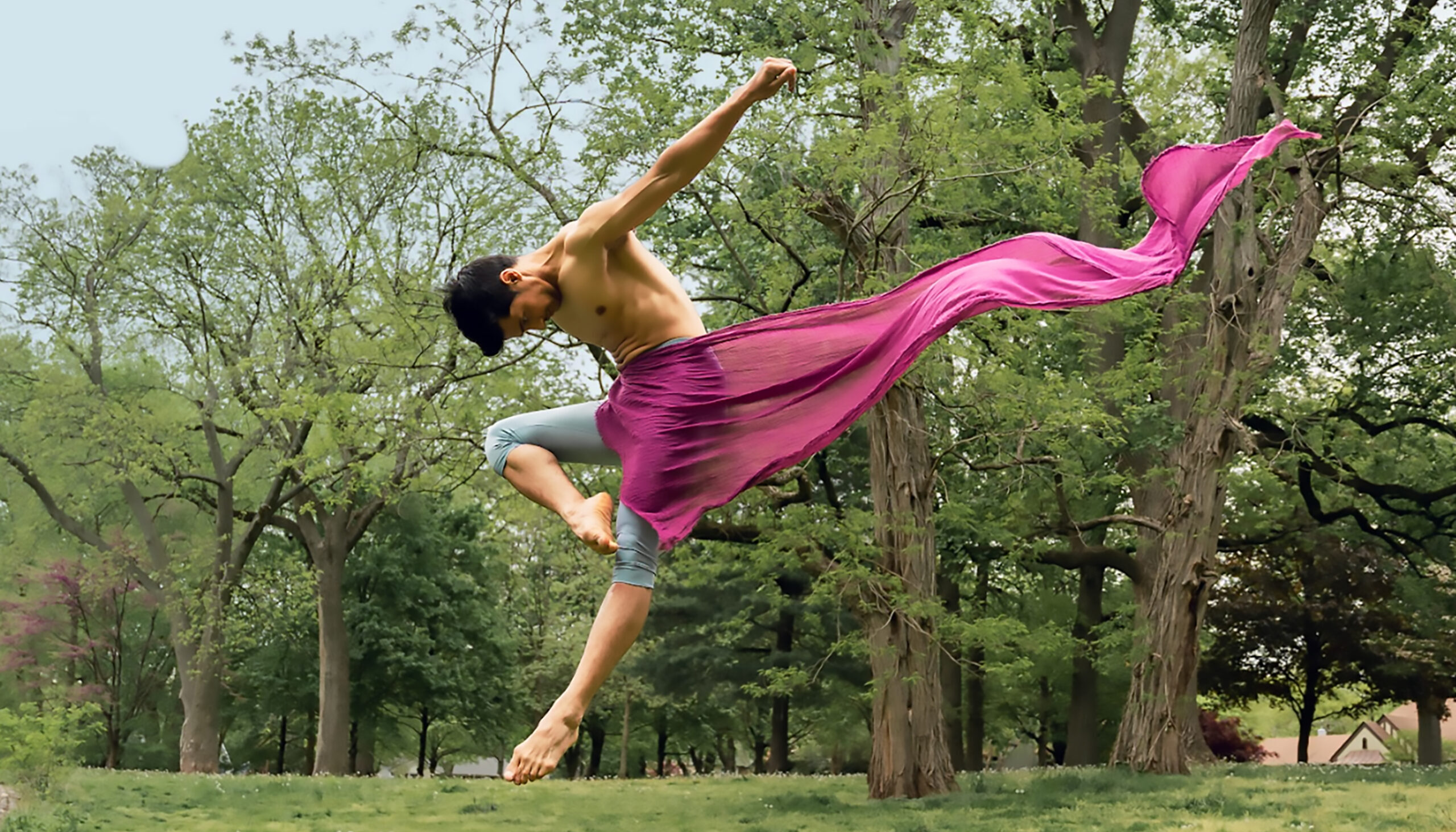 Manish Chauhan flies through the air outside in a stylized jump in retire devant, his body pitched forward and arms thrown back, a magenta scarf flowing behind him as it wraps around his front.