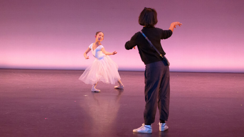 Georné Aucoin is shown from the back coaching a young dancer onstage. She wears s black shirt, gray sweatpants and white sneakers, with a small purse slung across her body. She lifts her right arm in a graceful position. In front of her, a teenage female dancer poses in a large fourth position lunge. She reaches her upper body forward slightly, with her arms moving toward her left in a softened pose. She wears a white short-sleeved leotard, a long white tutu, pink tights and pointe shoes, and smiles totwards the downstage left corner.