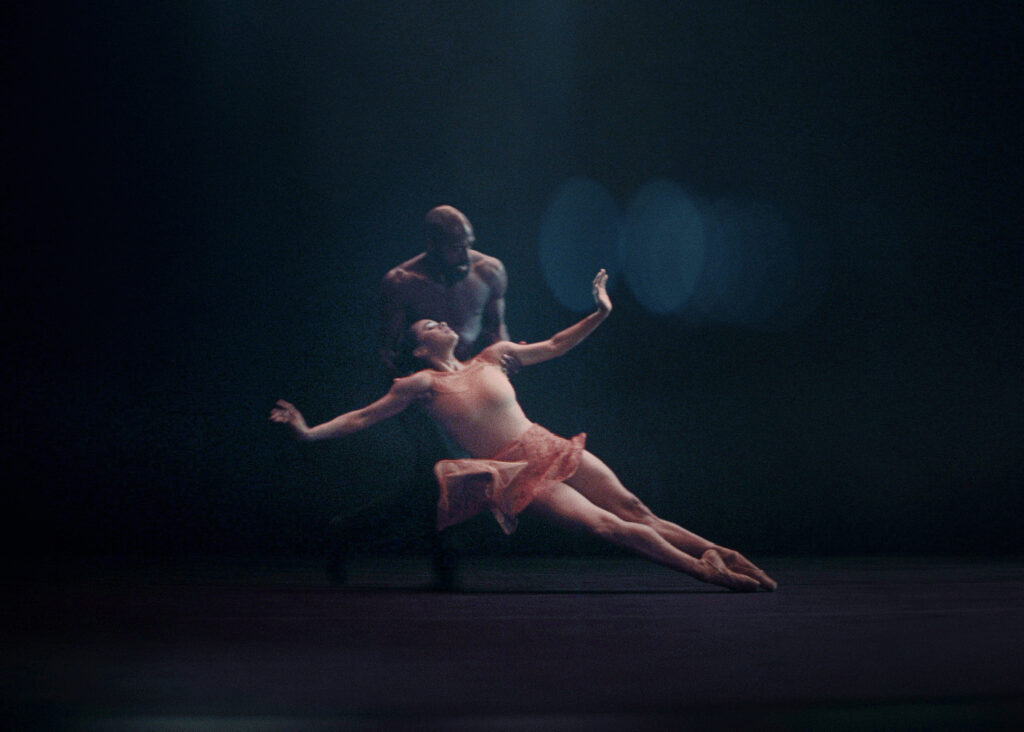 Misty Copeland, in a short pink dance dress, leans back, stretches her arms out, and crosses her legs in sus-sous. Babatunji Johnson, shirtless and in black pants, catches her under the armpits and starts to push her across the floor. They dance on a black floor and in front of a black backdrop.
