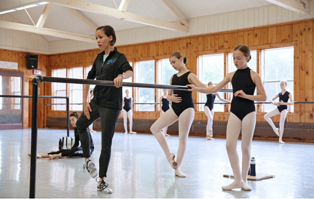 A female ballet instructor stands at a barre and demonstrates a coupé position with her right foot. A group of teenage female ballet studnets in black leotards, pink tights and pink ballet slippers watch and listen, with some copying her position. Cameron-Martin wears tight-fitting black pants, a zip-up athletic sweatshirt and white and black sneakers. The class is held in a large dance studio with wood-paneled walls and large windows.