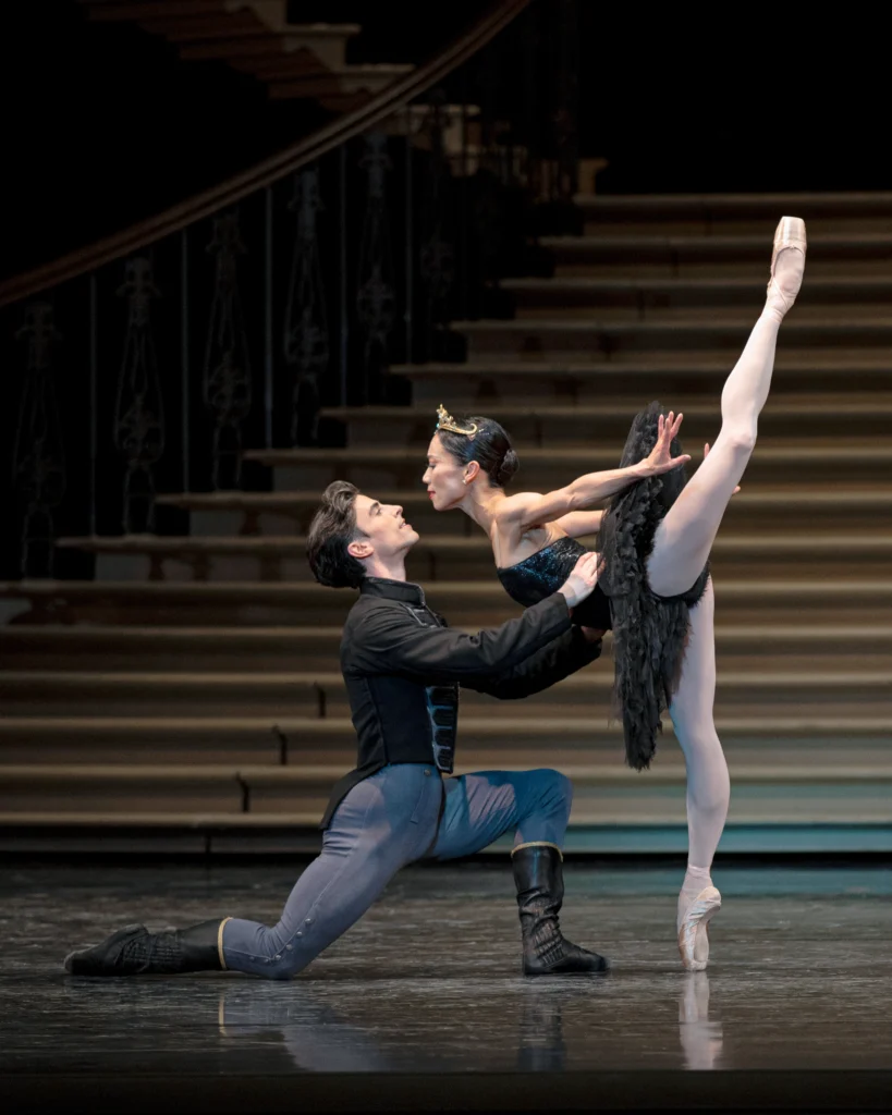 Frances Chung and Joseph Walsh perform the Black Swan pas de deux onstage during a performance. Walsh, in a black tunic, gray pants and black boots, kneels in profile on his left knee and holds Chung's waist. Chung, in a black tutu and crown, does a penché on pointe with her left leg in attitude and reaches her arms slightly back. They look at each other, their faces close to touching.