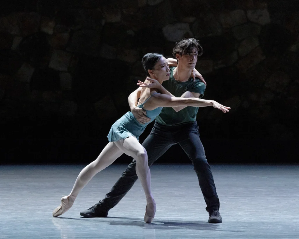 Frances Chung and Joseph Walsh dance a pas de deux onstage during a performance. Chung, in a blue leotard and skirt, lunges forward on pointe, reaching her right arm out as Walsh holds her around the waist and across her right shoulder. Walsh wears dark pants, sneakers and a green short-sleeved shirt. They both look towards downstage left with intensity.