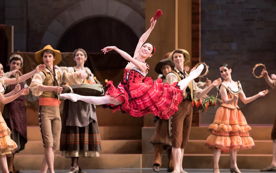 Francisc Chung, in pointe shoes and wearing a red Spanish-style dress with a knee-length, layered skirt, does a large leap with her back leg in attitude. She smiles triumphantly and holds her arms in third position, holding a red fan in her right hand. Other dancers stand behind her onstage wearing various Spanish peasant costumes. They watch her with big smiles, clapping to the music or banging tambourines.