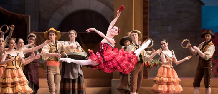 Francisc Chung, in pointe shoes and wearing a red Spanish-style dress with a knee-length, layered skirt, does a large leap with her back leg in attitude. She smiles triumphantly and holds her arms in third position, holding a red fan in her right hand. Other dancers stand behind her onstage wearing various Spanish peasant costumes. They watch her with big smiles, clapping to the music or banging tambourines.