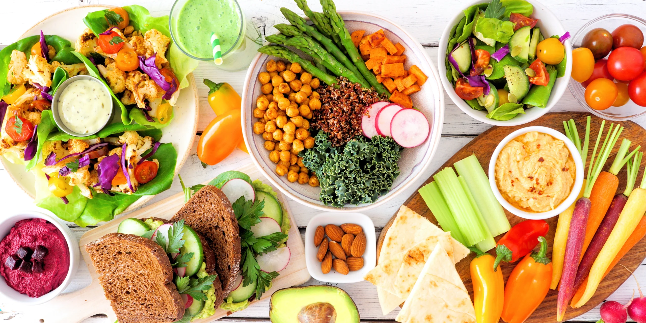 Platters and bowls of plant-based foods are shown from above on a white-washed table. They include salads, vegetarian sandwiches, and dips.
