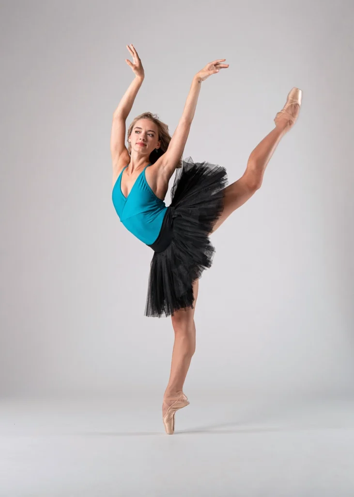 Grace Rookstool balances in back attitude on pointe. Her arms are raised in a soft V similar to Swan Lake. Her blonde hair is loose behind her shoulders. She wears a black practice tutu over a turquoise leotard.
