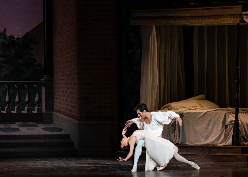 Yuka Oba-Muschiana and Josué Justiz perform a passionate pas de deux during a ballet performance of Romeo and Juliet. They perform in front of a large set with a canopy bed upstage left and a stone balcony on stage right. Justiz kneels down and Oba-Musciana lies across his right thigh, arching back as both she and Justiz open their arms in second. She tucks her left foot underneath her and extends her right leg out. She wears a light-colored, knee-length nightgown, tights and pointe shoes, while Justiz wears a white peasant blouse, tights, and ballet slippers.