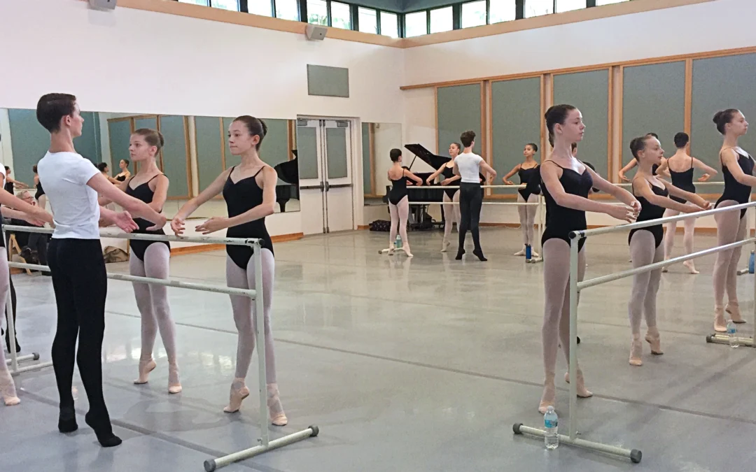 In a large dance studio, male a female ballet students stand at portable barres and balance in first position relevé. They face the barre and hold their arms in first position. The boys wear white t-shirts, black tights and black ballet slippers; the female students wear black leotards, pink tights, and pink ballet slippers with ribbons tied around the ankles. A grand piano can be seen in the far corner.