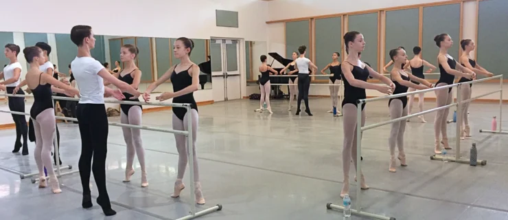 In a large dance studio, male a female ballet students stand at portable barres and balance in first position relevé. They face the barre and hold their arms in first position. The boys wear white t-shirts, black tights and black ballet slippers; the female students wear black leotards, pink tights, and pink ballet slippers with ribbons tied around the ankles. A grand piano can be seen in the far corner.