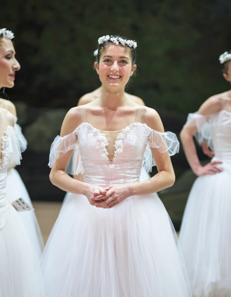 Federica Bona smiles towards the camera while backstage during a ballet performance. Sh wears a white Romantic-style tutu with filmy off-the-shoulder sleeves and a crown of white flowers in her dark hair, which is pulled back into a bun. She is shown standing from the thighs up, clasping her hands in front of her.