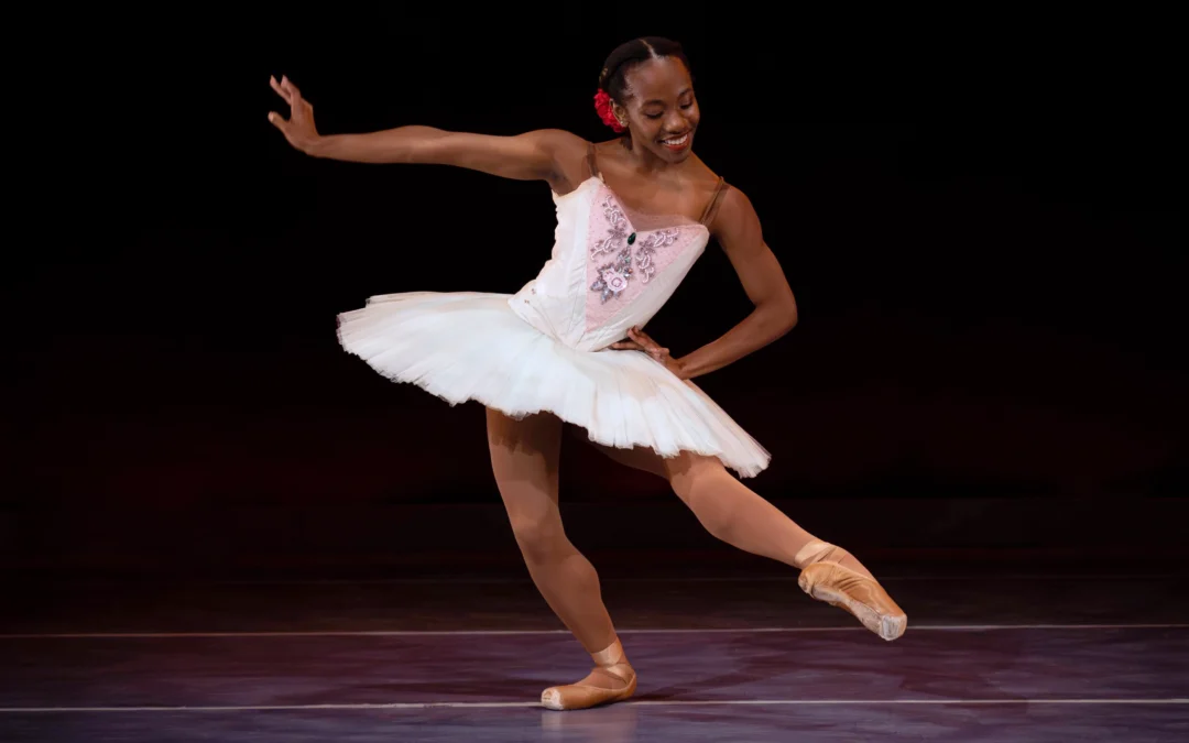 Marja Miller, who wears a white tutu with pink and floral trim on the bodice, brown tights and pointe shoes, and a red rose on the right side of her low bun, dances on stage in front of a dark backdrop. She pliés on her right leg and brushes her left leg into degagé devant in effacé. She extends her right arm to the side, flexing her wrist, puts her left hand on her hip, and looks down at her pointed foot with a smile.