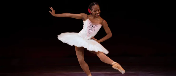 Marja Miller, who wears a white tutu with pink and floral trim on the bodice, brown tights and pointe shoes, and a red rose on the right side of her low bun, dances on stage in front of a dark backdrop. She pliés on her right leg and brushes her left leg into degagé devant in effacé. She extends her right arm to the side, flexing her wrist, puts her left hand on her hip, and looks down at her pointed foot with a smile.