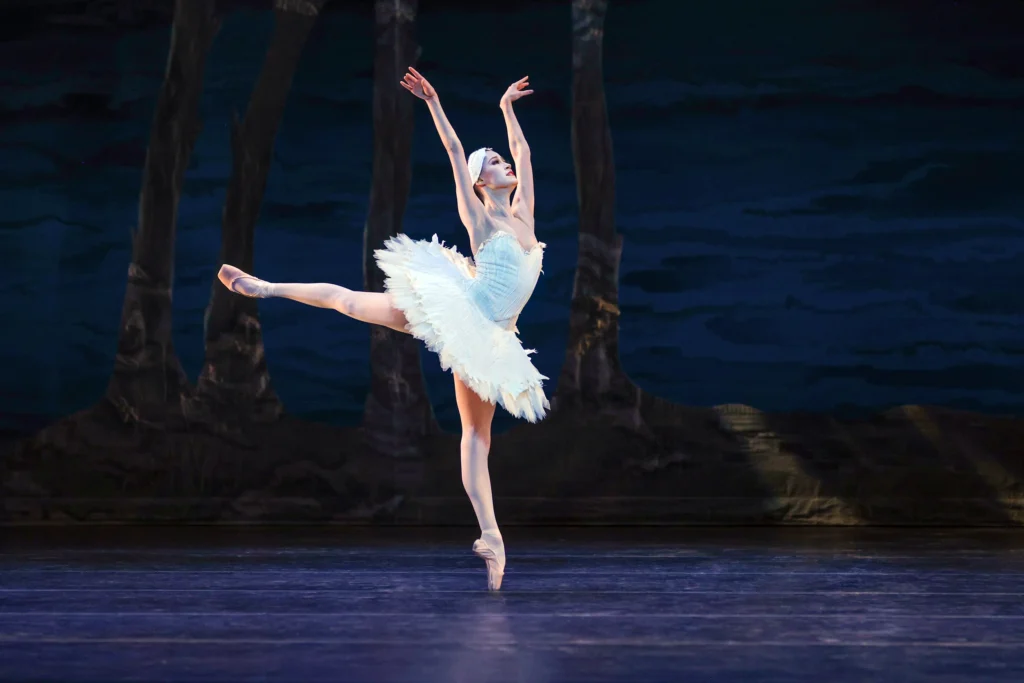 Jindallae Bernard balances in a clean first arabesque, arms high by her head. She wears a feathery white tutu and headpiece, pink tights, and pointe shoes.