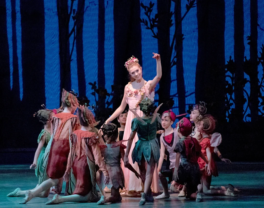 During a performance of A Midsummer Night's Dream, Miriam Miller, wearing a long pink gown and floral headpiece, is surrounded by a circle of small children. They face her, with most on one knee and one child standing in front of Miller as she acknowledges them. The little dancers wear various costumes like bugs and butterflies. They dance in front of a backdrop portraying a dark forest against a dark blue background.
