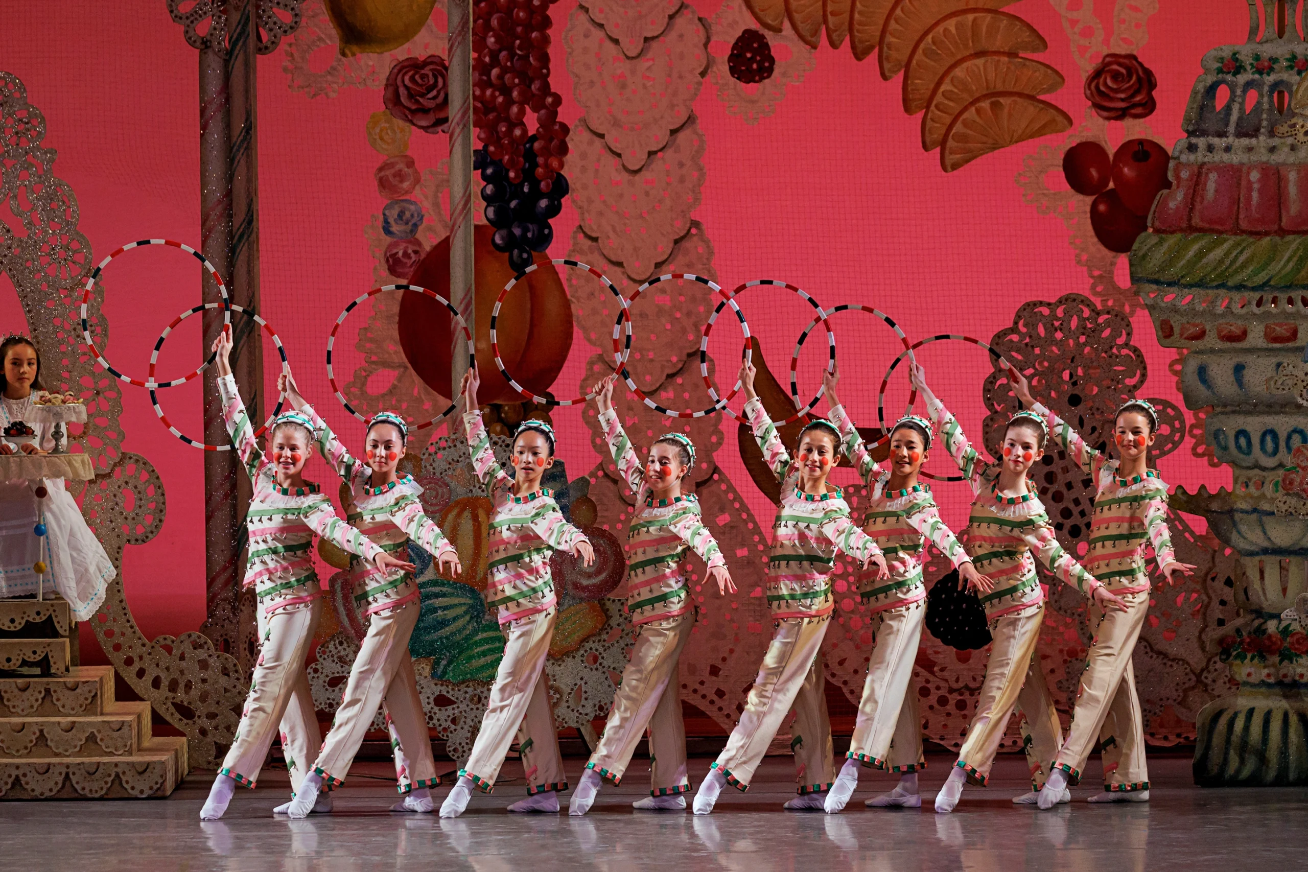 During a performance of The Nutcracker, a group of pre-teen female dancers stand in a line with their left leg in tendu devant croise. They wear costumes of white pants, tunics and caps with pink and green stripes and matching pom-pom embellishments. They wear red circles on their cheeks and hold a candy-cane striped hoop in their raised right hand. They poses in front of an elaborate set depicting a land of sweets, with tiers of candy and sugar.