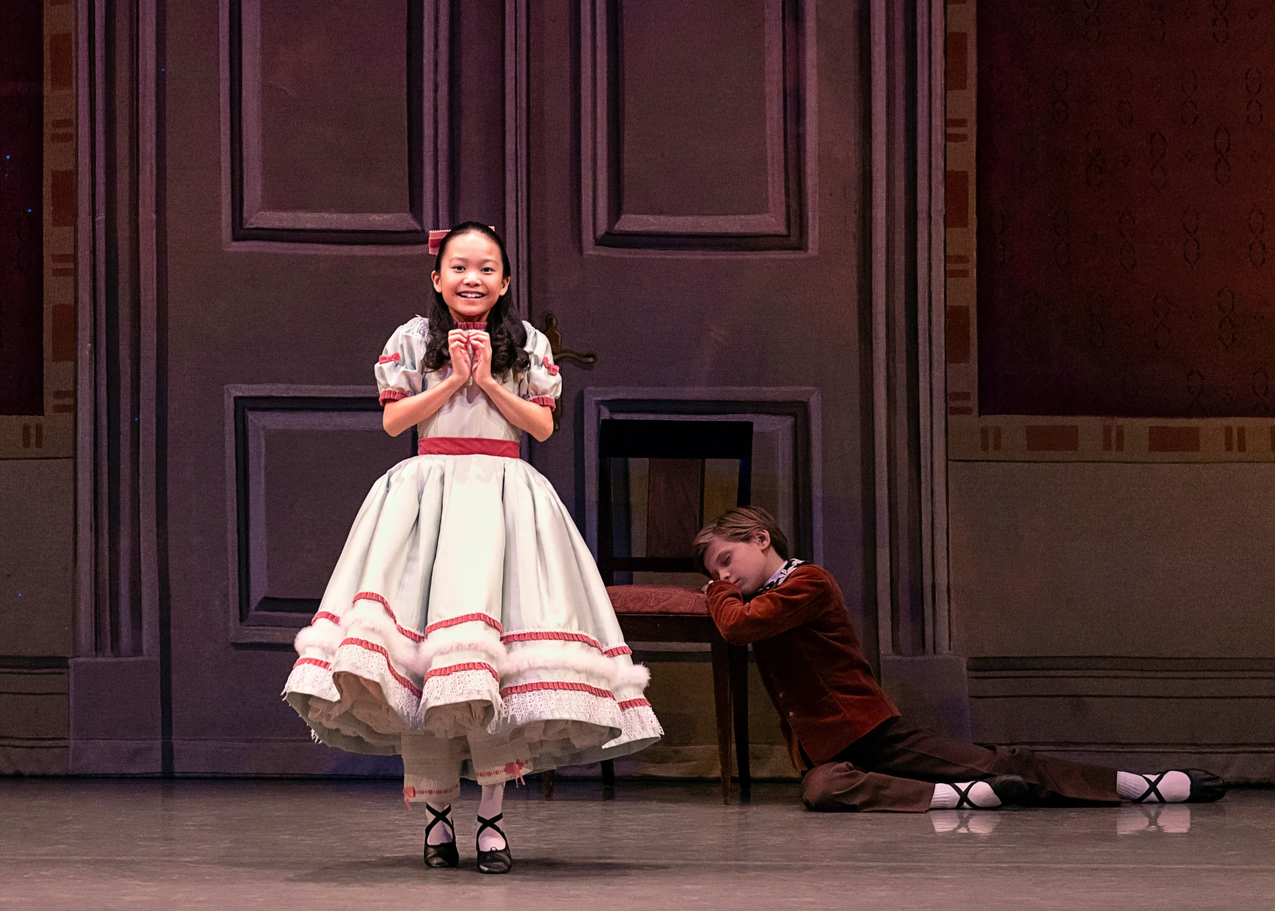 During a performance of The Nutcracker, a young dancer stands on her tiptoes and holds her hands together under her chin in excitement. She wears a long, full silver Victorian dress with a pink ribbon around the waist and pink trimmings around the skirt and puffed sleeves, and a big pink bow in her hair. Behind her, a young boy dancer sits on the floor with his legs out to his left and rests his head on his hands on the seat of a chair, napping. They poses in front of a giant parlor door.