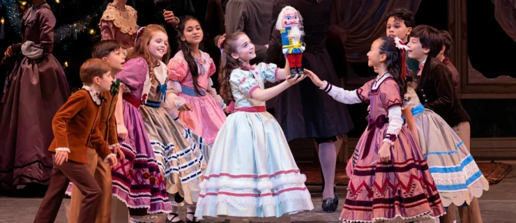 During a performance of The Nutcracker, a young girl dancer stands center stage and holds up a nutcracker doll, surrounded by other child dancers. She wears a silver Victorian style dress with a long, full skirt, short puffed sleeves, and pink trimmings along the skirt, sleeves and waist. The girls surrounding her also wear long dresses in various colors, and the boys wear pants and buttoned jackets in different shades of brown. Behind them, adults in period costume mingle during the in front of a large Christmas tree.