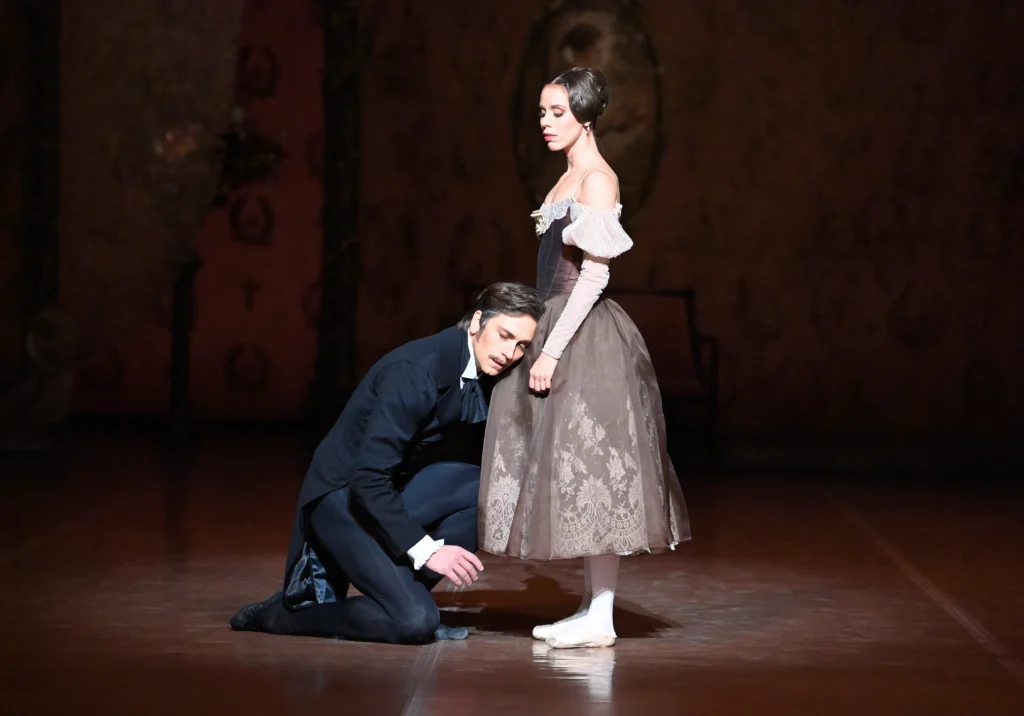 During a performance of the ballet Onegin, Friedemann Vogel kneels on the ground and rests his head despairingly on the skirt of Elisa Badenes. He wears a black jacket with tails, black tights and black ballet slippers. Badenes looks down at him, her arms at her side. She wears a brown dress with a long, full skirt and puffed, off-the-shoulder sleeves. Her hair is worn in a Romantic-style bun.