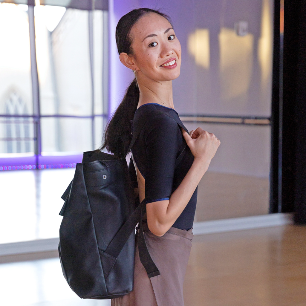 A profile shot of Ryoko Tanaka smiling over her right shoulder at the camera. She is wearing a purple leotard and mauve skirt with a black backpack.