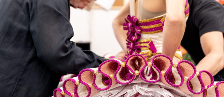 A close-up photo shows a female dancer being fitted for a ruffly gold and magenta pancake tutu.