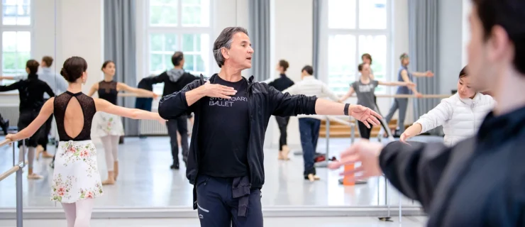 Tamas Detrich teaches class in a large ballet studio full of professional dancers. He holds his left arm out in second position and looks out over it, while touching his chest with his right hand to demonstrate an open feeling. He wears a black shirt with the words Stuttgart Ballet on it in white, a dark, unzipped hoodie sweatshirt, and blue sweatpants.